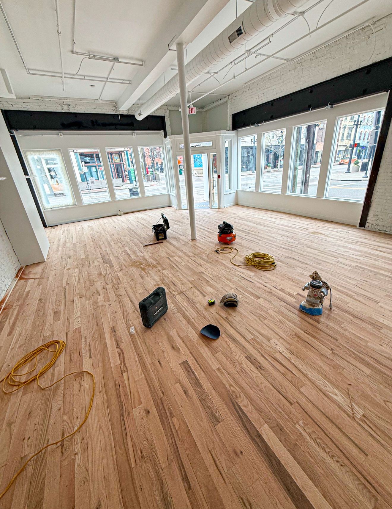 Empty room with wooden flooring, power tools, and large windows overlooking a street.